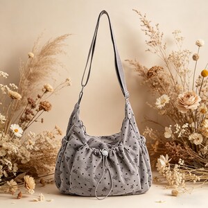 May include: A gray shoulder bag with a repeating pattern of small hearts and bear shapes. The bag has a drawstring closure and a long, adjustable strap. The background features dried flowers and a neutral backdrop.