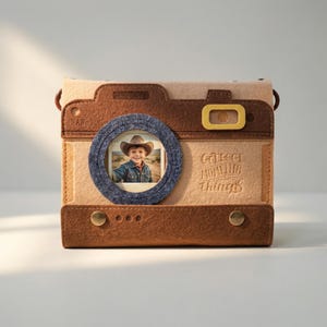 May include: A tan and brown felt photo album shaped like a camera, with a circular photo frame and the text "Collect Moments, Not Things." The album features a small photo of a child wearing a cowboy hat. It has a brown strap and gold-coloured button details.