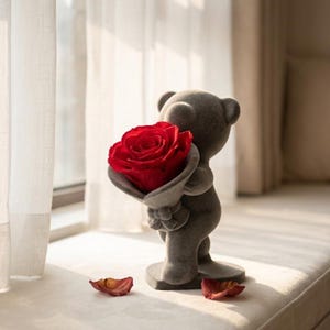 Puede incluir: Una figura de oso de peluche gris sosteniendo una gran rosa roja. El oso está de pie sobre un pedestal, con dos pétalos de rosa caídos cerca. La rosa está en plena floración, con una textura aterciopelada.