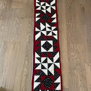 May include: A long, rectangular table runner featuring a patchwork design of red, white, and black fabric. The pattern consists of repeating star and square blocks with a small white star print on black fabric and a red and black checkered border.