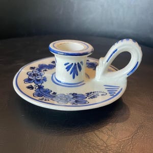 May include: A white ceramic candle holder with a circular base and a side handle. The piece features hand-painted blue floral patterns and leaf motifs around the central candle cup, with blue line accents on the rim and handle edges.