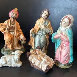 May include: A nativity set featuring five ceramic figurines: a kneeling figure in an orange robe and pink turban, a kneeling figure in a tan and green robe, a kneeling figure in a pink dress and blue head covering, a baby in a manger, and a white lamb.
