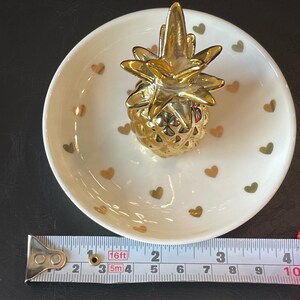 May include: A white ceramic trinket dish featuring a gold-coloured pineapple figurine in the centre and small gold heart patterns on the surface. A measuring tape placed at the base shows the dish has a diameter of approximately 10 cm.