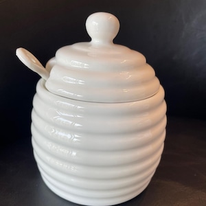 Puede incluir: Un tarro de miel de cerámica blanca con una textura acanalada inspirada en una colmena. El recipiente cuenta con una tapa a juego con un pequeño pomo y una muesca lateral para una cuchara. Una pequeña cuchara de cerámica blanca está insertada.
