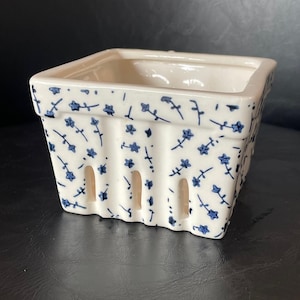 Puede incluir: Una cesta cuadrada de cerámica para bayas con esmalte blanco roto, decorada con un patrón pintado a mano de pequeñas flores y enredaderas azules. El recipiente tiene un diseño acanalado con aberturas verticales en la base de sus cuatro lados.