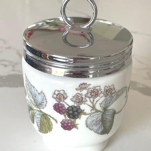 May include: A white ceramic jar with a polished silver-tone metal lid featuring a circular handle. The body of the jar is decorated with a botanical illustration of green leaves, white flowers, and clusters of purple and black blackberries.