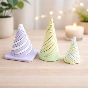 May include: Three decorative cone-shaped objects in pastel shades. The largest is lavender with white stripes, the middle one is green and yellow, and the smallest is white and green. They are displayed on a light wooden surface.