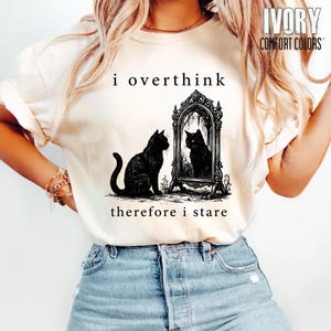 Puede incluir: Camiseta crema Comfort Colors con el texto "i overthink therefore i stare". El gráfico presenta un gato negro mirando su reflejo en un espejo decorativo.