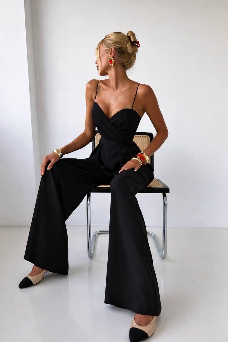 Op de afbeelding: Een zwarte mouwloze jumpsuit met een hartvormige halslijn, een overslagtop, dunne spaghettibandjes en een broek met wijde pijpen. Het kledingstuk wordt gecombineerd met goudkleurige armbanden, rode oorbellen en beige met zwarte schoenen met neuskap.
