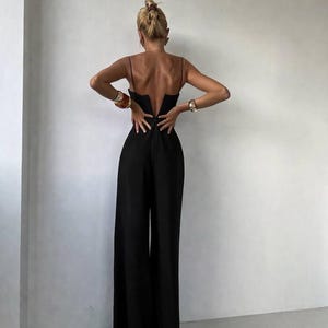 Op de afbeelding: Een zwarte jumpsuit met een diepe V-vormige uitsnede op de rug, dunne spaghettibandjes en een broek met wijde pijpen. Het kledingstuk valt tot op de grond en heeft een aansluitend lijfje dat overgaat in een losvallend, vloeiend silhouet.