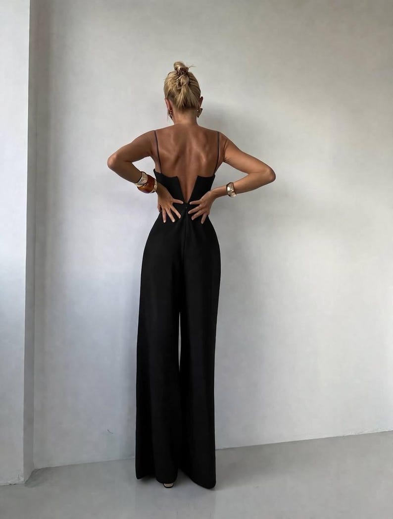 Op de afbeelding: Een zwarte jumpsuit met een diepe V-vormige uitsnede op de rug, dunne spaghettibandjes en een broek met wijde pijpen. Het kledingstuk valt tot op de grond en heeft een aansluitend lijfje dat overgaat in een losvallend, vloeiend silhouet.