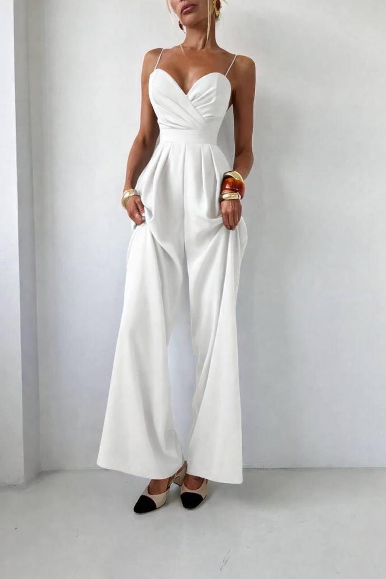 Op de afbeelding: Een effen witte jumpsuit met een hartvormige halslijn, dunne spaghettibandjes en een getailleerd lijfje met een geplooide taille. Het kledingstuk heeft een broek met wijde pijpen die tot op de grond valt, wat zorgt voor een strak en gestructureerd silhouet.