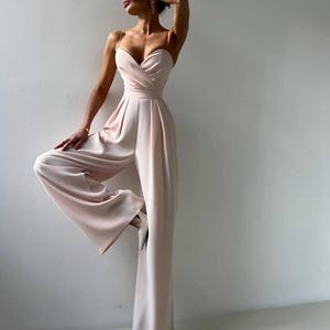 Op de afbeelding: Een mouwloze jumpsuit in lichtroze met een hartvormige halslijn, een overslagtop, dunne spaghettibandjes en een broek met wijde pijpen. Het kledingstuk heeft een getailleerde pasvorm en een gladde, effen afwerking van de stof.