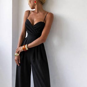 Op de afbeelding: Een zwarte mouwloze jumpsuit met een hartvormige halslijn, gedrapeerde details op het lijfje, dunne spaghettibandjes en een broek met wijde pijpen. Het kledingstuk is gecombineerd met goudkleurige armbanden, een grote barnsteenkleurige armband en oorbellen.