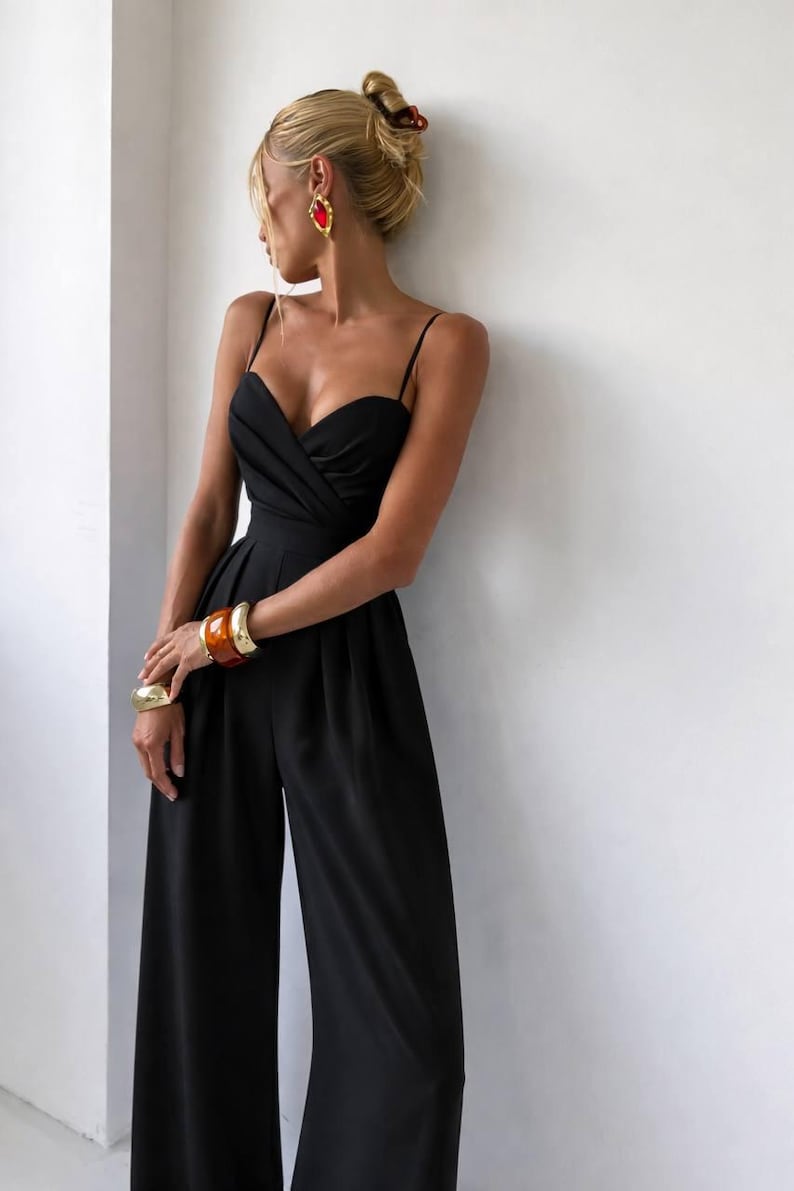 Op de afbeelding: Een zwarte mouwloze jumpsuit met een hartvormige halslijn, gedrapeerde details op het lijfje, dunne spaghettibandjes en een broek met wijde pijpen. Het kledingstuk is gecombineerd met goudkleurige armbanden, een grote barnsteenkleurige armband en oorbellen.