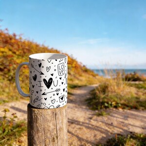 Op de afbeelding: Witte keramische mok met een zwart doodle-ontwerp van harten, pijlen en de woorden "I love you" en "love". De mok staat op een houten paal met een strand en blauwe lucht op de achtergrond.