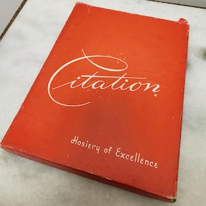 May include: A rectangular orange box with the word Citation written in a large white cursive script across the top half. Below the script, the text Hosiery of Excellence is printed in a smaller white sans-serif font on the front cover.