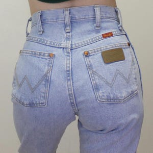 May include: Light blue high-waisted denim jeans featuring a classic five-pocket design, copper-toned rivets, and a tan leather patch on the back pocket with the Wrangler brand name. The back pockets display a signature W-shaped stitching pattern.