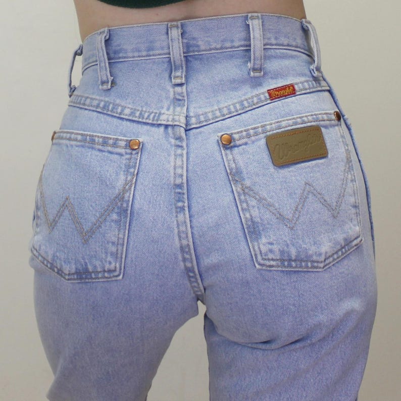 May include: Light blue high-waisted denim jeans featuring a classic five-pocket design, copper-toned rivets, and a tan leather patch on the back pocket with the Wrangler brand name. The back pockets display a signature W-shaped stitching pattern.
