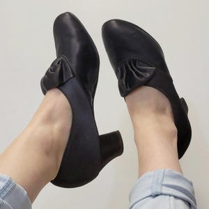 May include: A pair of black leather shoes with a rounded toe and a small heel. Each shoe features a decorative bow on the top. The shoes are designed with an open side and a strap around the ankle.