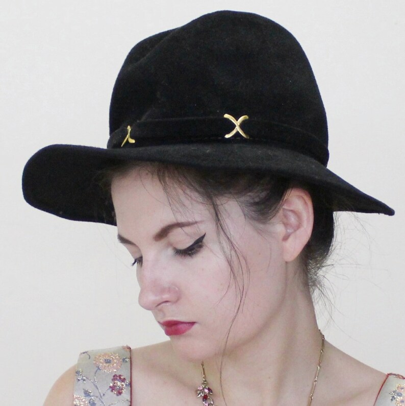 Puede incluir: Un sombrero fedora de fieltro negro con un dise&ntilde;o en X dorado en la banda. El sombrero lo lleva una persona con el pelo oscuro y una expresi&oacute;n neutral.