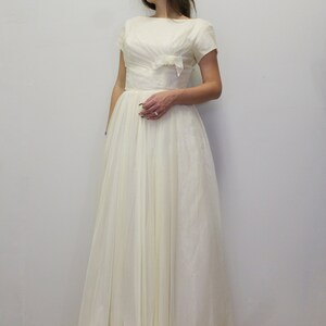 May include: A white vintage wedding dress with a bateau neckline, short sleeves, and a fitted bodice with a bow detail. The skirt is full and flowing.