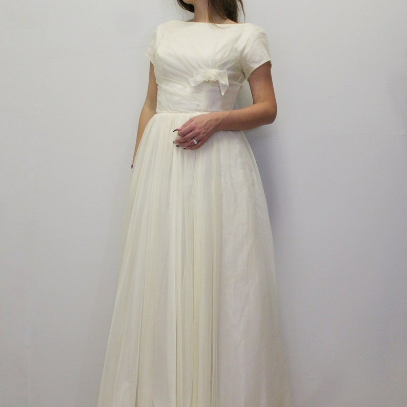 May include: A white vintage wedding dress with a bateau neckline, short sleeves, and a fitted bodice with a bow detail. The skirt is full and flowing.