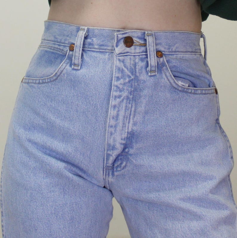 May include: High-waisted light blue denim jeans featuring a classic five-pocket design, a metal button closure at the waist, belt loops, and copper-toned rivets. The fabric has a faded wash appearance with visible stitching along the seams.