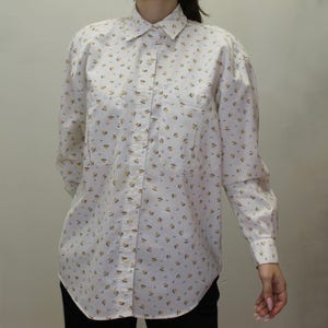 May include: A long-sleeve button-down shirt in white featuring a repeating pattern of small yellow flowers with green stems. The shirt includes a standard collar, a front button placket, and two chest pockets with button closures on the front.