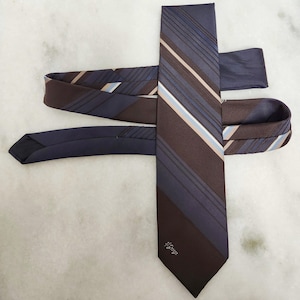 May include: A dark brown and navy blue striped tie with light blue and tan accents. The tie has a diagonal stripe pattern and a small floral design near the tip. The tie is made of a smooth, likely silk, material.