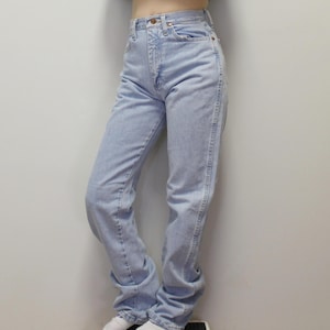 May include: High-waisted light wash denim jeans featuring a classic five-pocket design, a metal button closure at the waist, and a straight-leg cut. The fabric displays a faded blue color with visible stitching along the seams and pockets.