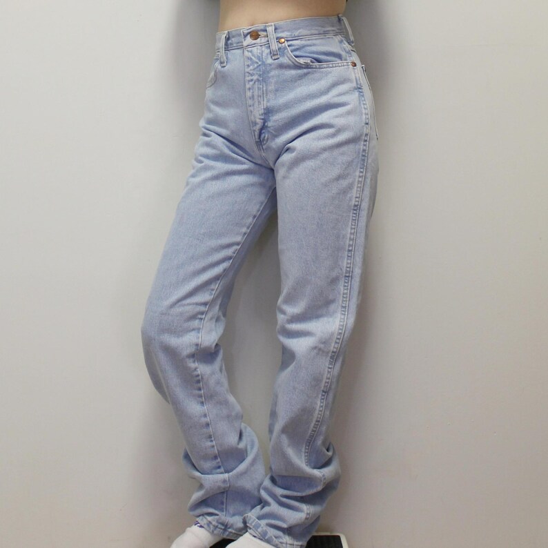 May include: High-waisted light wash denim jeans featuring a classic five-pocket design, a metal button closure at the waist, and a straight-leg cut. The fabric displays a faded blue color with visible stitching along the seams and pockets.