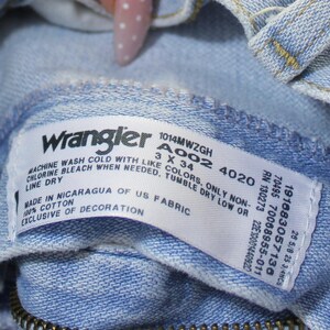 May include: A light blue denim garment featuring a white interior tag with the Wrangler logo, model number 1014MWZGH, size 3 x 34, and care instructions. The tag specifies 100% cotton, made in Nicaragua of US fabric, and exclusive of decoration.