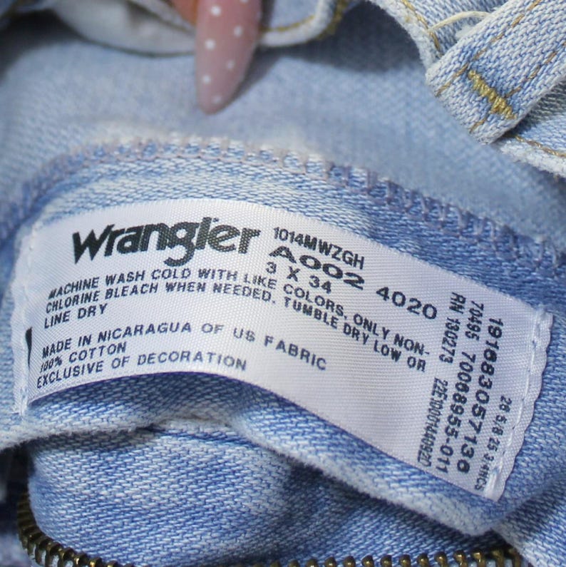 May include: A light blue denim garment featuring a white interior tag with the Wrangler logo, model number 1014MWZGH, size 3 x 34, and care instructions. The tag specifies 100% cotton, made in Nicaragua of US fabric, and exclusive of decoration.
