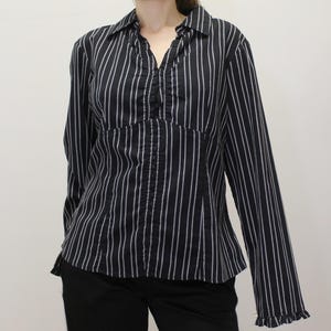 Puede incluir: Una camisa negra de manga larga con finas rayas verticales blancas, cuello con tapeta con volantes y cierre frontal de botones. La prenda presenta un cuerpo entallado con costuras verticales y puños con volantes en cada manga.