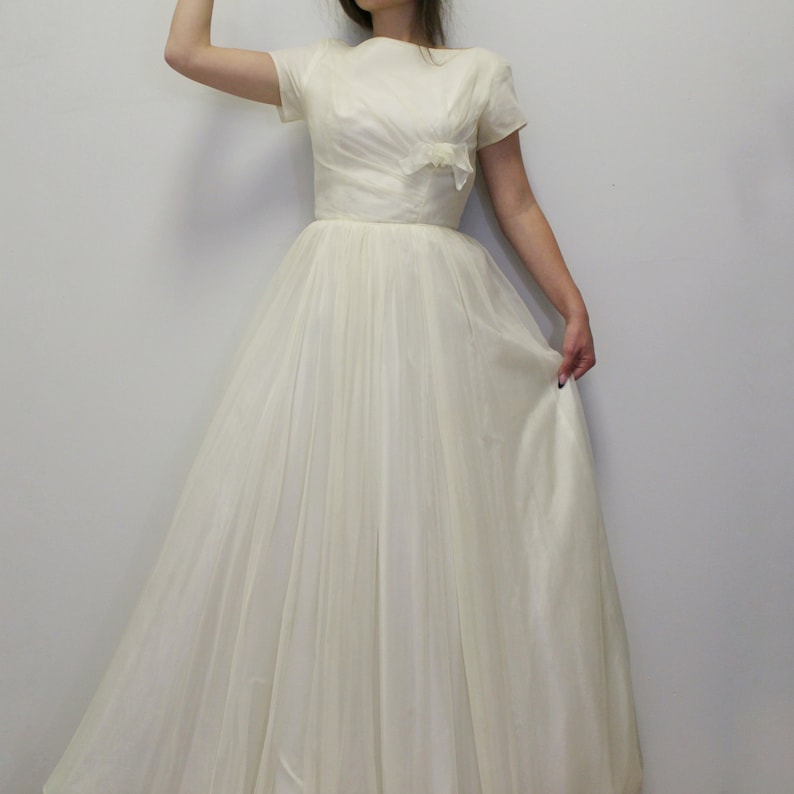 May include: A white vintage wedding dress with a fitted bodice and a full skirt. The dress has short sleeves and a bateau neckline.