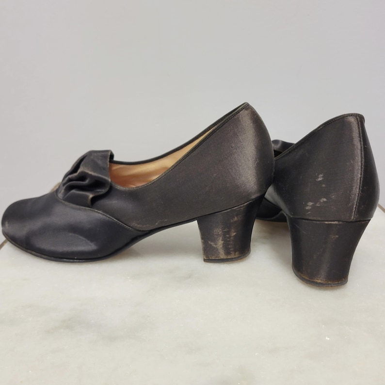 May include: A pair of vintage dark brown satin pumps with a small heel. The shoes feature a decorative ruffle detail on the vamp. The heels show signs of wear. The shoes are likely from the early to mid-20th century.