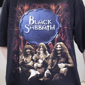 May include: Black Sabbath band t-shirt featuring a black and white image of the band members in front of a dark forest background. The band's name is written in white letters on a blue circle.