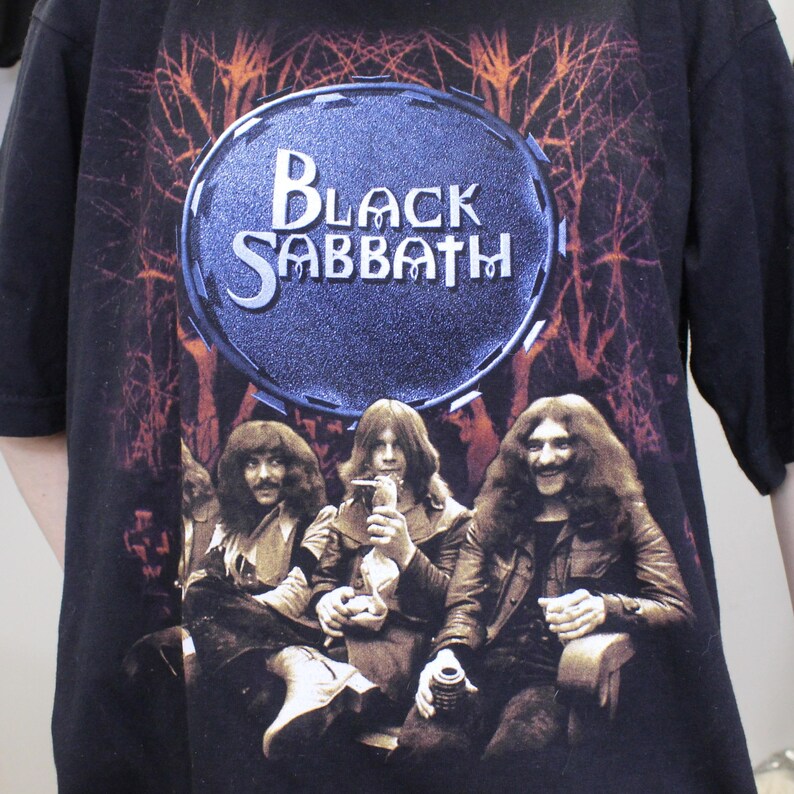 May include: Black Sabbath band t-shirt featuring a black and white image of the band members in front of a dark forest background. The band's name is written in white letters on a blue circle.