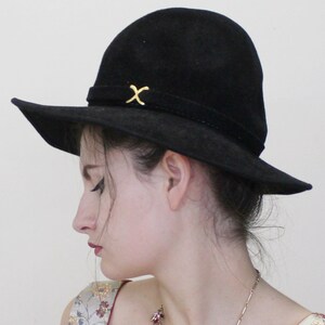 Puede incluir: Un sombrero negro de ala ancha con un detalle en X dorado en la banda. El sombrero est&aacute; hecho de un material suave, similar al ante.