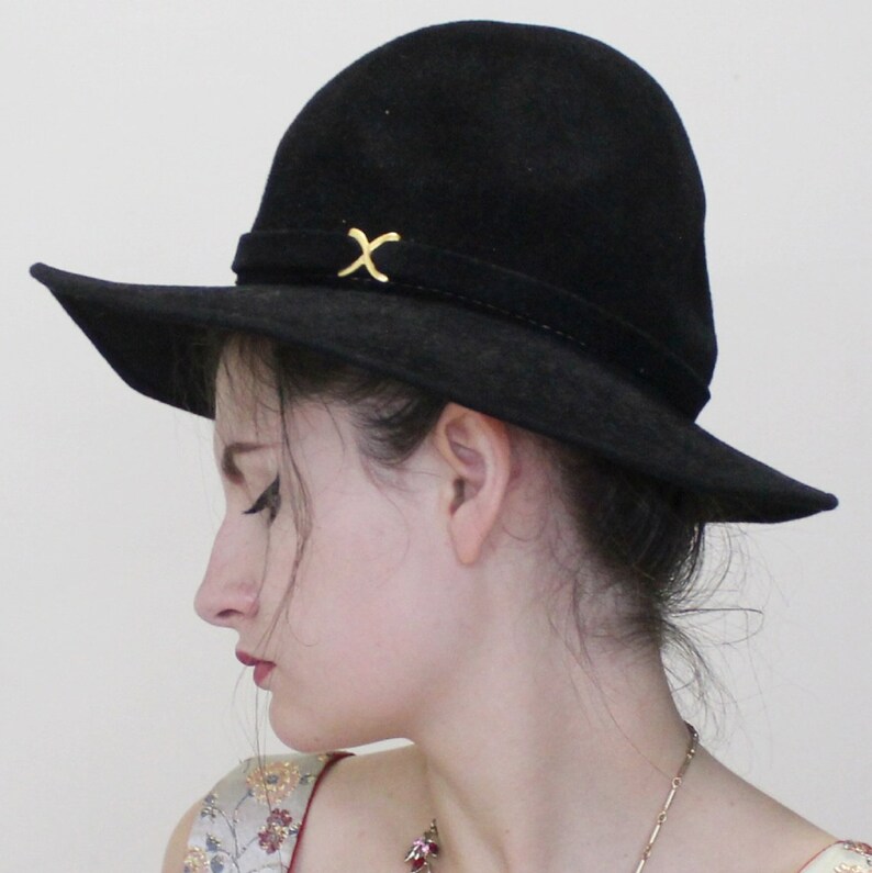 Puede incluir: Un sombrero negro de ala ancha con un detalle en X dorado en la banda. El sombrero est&aacute; hecho de un material suave, similar al ante.