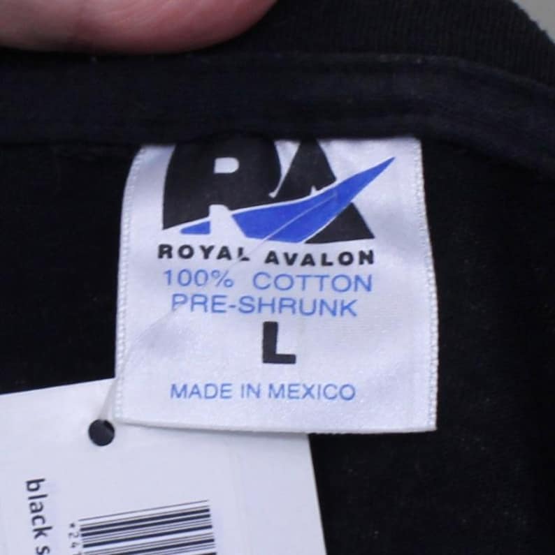May include: A white tag with black text that reads "ROYAL AVALON 100% COTTON PRE-SHRUNK L MADE IN MEXICO". The tag is attached to a black shirt.