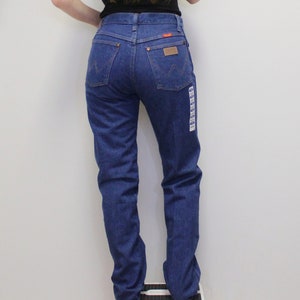 May include: A pair of blue denim jeans with a high waist and a straight leg. The jeans have a brown leather patch on the back with the Wrangler logo.