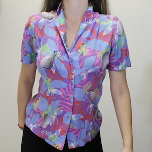 Peut inclure: Une chemise à manches courtes boutonnée avec un motif floral vibrant. La chemise présente une base violette avec des motifs de feuilles et de poires bleues, vertes et oranges. Le col et les boutons sont visibles.