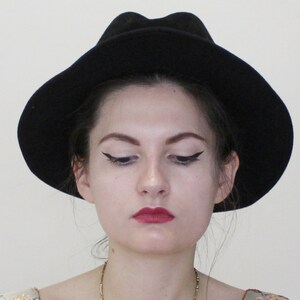 Puede incluir: Una mujer con cabello oscuro lleva un sombrero fedora negro. Tiene delineador de ojos oscuro y l&aacute;piz labial rojo.