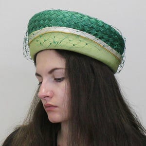 May include: A green and light green pillbox hat with a white ribbon and a green netting veil.