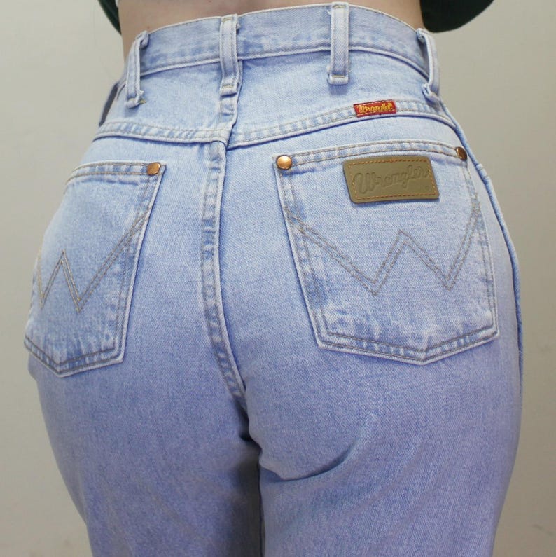 May include: Light blue high-waisted denim jeans featuring a classic five-pocket design, copper-toned rivets, and a tan leather patch on the back pocket with the Wrangler brand name. The back pockets display a signature W-shaped stitching pattern.