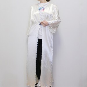 May include: A white satin robe with a scalloped trim. The robe has a long, flowing silhouette and a tie closure at the waist.