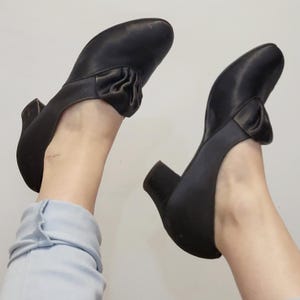 May include: A pair of black leather shoes with a low heel. Each shoe features a decorative bow detail on the top. The shoes are viewed from below.