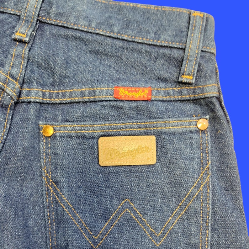 May include: A pair of blue denim jeans with a brown leather patch that says "Wrangler" on the back pocket. The jeans have a red leather patch that says "Wrangler" above the back pocket.